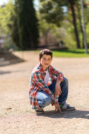 A young Caucasian boy with short dark hair, wearing a plaid shirt, jeans, and sneakers, squats on a gravel path in a sunlit park, looking curious.の写真素材
