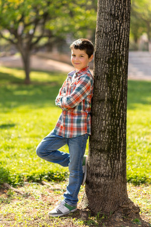 A young Caucasian boy with short dark hair, wearing a plaid shirt and jeans, leans against a tree in a sunlit park, smiling confidently.の写真素材