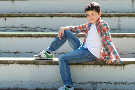 A young Caucasian boy with short dark hair, wearing a plaid shirt, jeans, and sneakers, sits casually on outdoor steps, smiling with a relaxed pose.の写真素材
