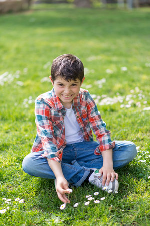 A young Caucasian boy with short dark hair, wearing a plaid shirt, jeans, and sneakers, sits cross-legged on grass in a park, picking daisies with a smile.の写真素材
