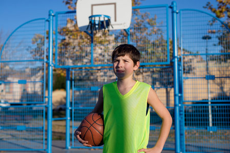 A young boy in a neon yellow jersey stands on an outdoor basketball court, holding a ball. He looks determined, with a metal hoop and blue fence behind him.の写真素材