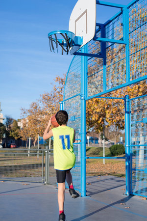 A young boy in a neon yellow jersey attempts a basketball shot at an outdoor court. The hoop is damaged, and autumn trees with golden leaves are visible.の写真素材