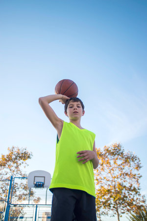 A young boy in a neon yellow jersey holds a basketball above his shoulder, standing confidently on an outdoor court. Blue sky and trees in the background.の写真素材