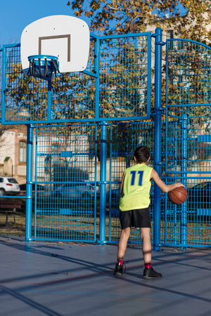 A young boy wearing a neon yellow jersey dribbles a basketball on an outdoor court. He faces a blue...