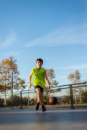 A young boy in a neon yellow jersey dribbles a basketball on an outdoor court. He is focused, moving forward, with trees and a clear sky in the background.の写真素材