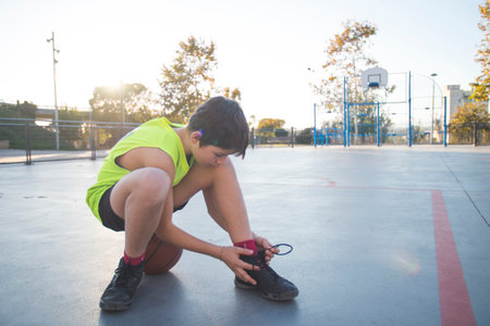 A young boy in a neon yellow jersey ties his shoe on a basketball court, sitting on a ball. The background features a hoop, fence, and trees at sunset.の写真素材