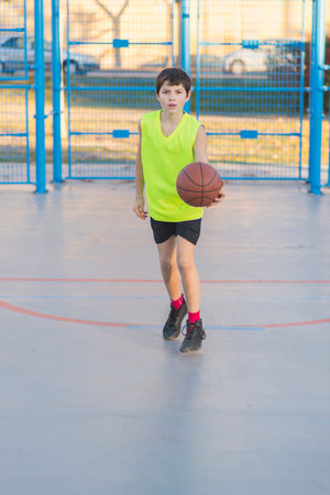 A young boy wearing a neon yellow jersey dribbles a basketball on an outdoor court. He is focused, moving forward, with autumn trees and mountains behind.の写真素材