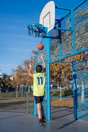 A young boy in a neon yellow jersey attempts a basketball shot at an outdoor court. The hoop is damaged, and autumn trees with golden leaves are visible.の写真素材