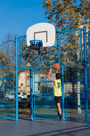 A young boy in a neon yellow jersey attempts a basketball shot at an outdoor court. The hoop is...