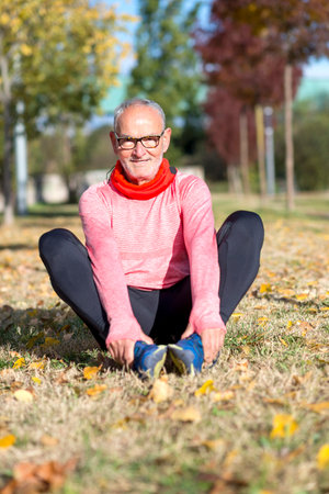Senior Caucasian man in sportswear sitting on grass in a park, stretching after a run, wearing glasses, pink long-sleeve shirt, black leggings.の写真素材