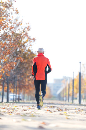 Senior Caucasian man jogging outdoors on a sunny day, wearing a pink long-sleeve top, black leggings, a red scarf, and sports shoes, viewed from behind.の写真素材