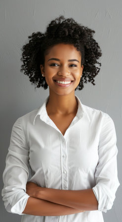 Confident young woman with natural curly hair, African descent, wearing a white shirt, smiling with arms crossed, standing against a gray wall, looking positive.の素材