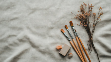 Five wooden-handle paintbrushes, soap, wool, and dried flowers arranged on textured light fabric. Minimal still life with natural and creative feel.の素材