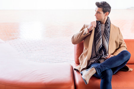 Caucasian young man sitting on a modern leather sofa, wearing a camel coat, jeans, and scarf. He...