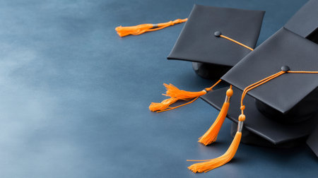 Close-up of black academic graduation caps with vibrant orange tassels placed on a blue textured background, symbolizing achievement and ceremony.の素材