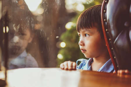 Kid looking out window. Adorable little caucasian kid boy stand still near window glass and looking to outside alone during bright sunny day. Little child reflecting bored and loneliness at homeの写真素材