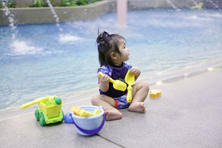 Asian baby girl playing with water toys by the pool. Little girl playing in outdoor swimming pool on summer vacation on tropical beach island. Child learning to swim in outdoor pool of luxury resort. Water toy for kidsの写真素材