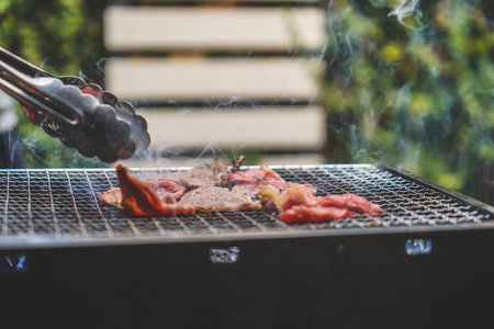 Close up view of pieces of sliced beef on charcoal grill at Outdoor party. Human's hands using meat tongs to grill Multiple slices of premium meat on the stovetop. Home made barbecue beef grill smoke.の写真素材