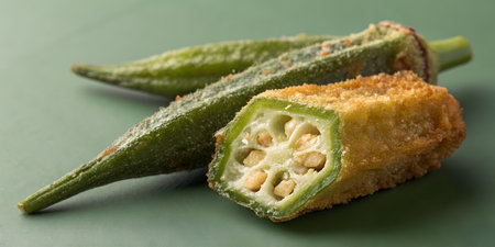 Fresh green okra vegetables prepared for eating. One pod is whole and the other cut showing internal seeds and slime. Coated in golden crunchy fried batter. Traditional southern side dish studio photograph against dark green background.の写真素材