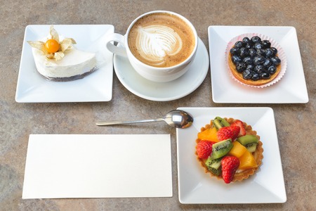 Different fruit cakes and cup of capuccino with blank card on table