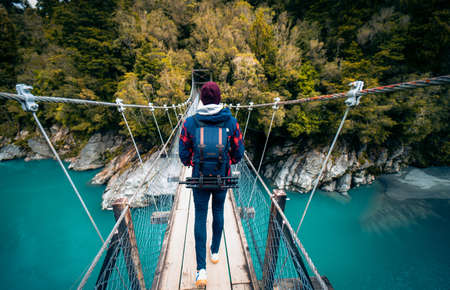 Hokitika Gorge, New Zealand, October 7, 2019: Beautiful panorama of a European man with a photographic backpack crossing a suspension bridge over a turuqesa blue water riverのeditorial素材