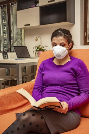 Corona Virus, Covid-19. Stay at home. Quarantine. Protection mask. A beautiful Caucasian woman forced to stay home to prevent virus infection, reads a book on the sofa to pass the time.の写真素材