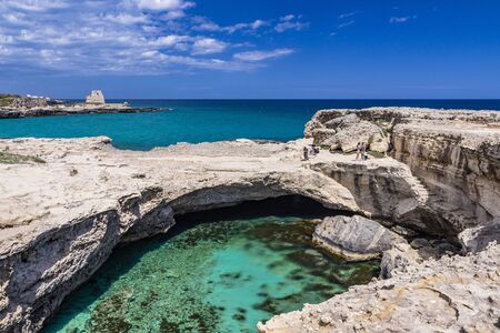 Archaeological site and tourist resort of Roca Vecchia, Puglia, Salento, Italy. Turquoise sea, clear blue sky, rocks, sun. The Cave of Poetry. Tourists sunbathe. The lookout tower in the background.の写真素材