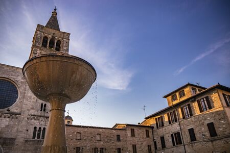 The Romanesque Church of San Michele in the medieval village of Bevagna. Umbria, Italy. The travertine facade, with the large rose window and the Gothic bell tower. The marble fountain. At sunsetの写真素材