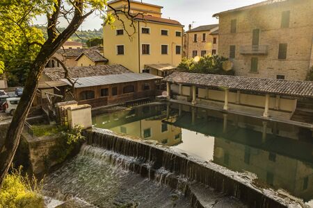 The ancient wooden wash house, on the river, in the medieval village of Bevagna. Umbria, Italy. The blue sky at sunset. Trees and vegetation. The reflection of the buildings on the water surface.の写真素材
