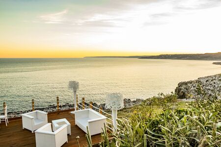 Italian restaurant with panoramic terrace, with spectacular view of the blue sea at sunset. In Puglia, Italy. Wooden terrace with curtains, armchairs, chairs, tables and lamps in white.の写真素材