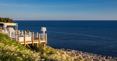 Italian restaurant with panoramic terrace, with spectacular view of the blue sea. A wonderful place in Puglia, Italy. Wooden terrace with curtains, armchairs, chairs, tables and lamps in white.の写真素材