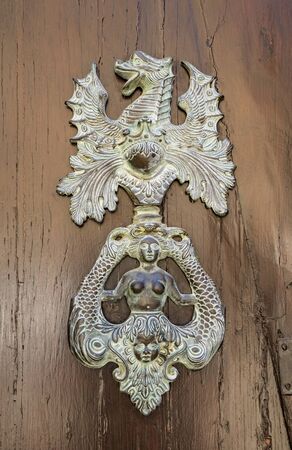 An antique metal knocker on a wooden door, which depicts a dragon and a mermaid.の写真素材