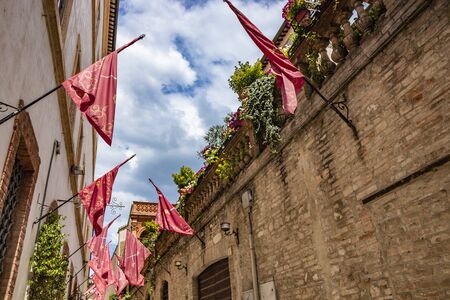 June 1, 2019 - Foligno, Perugia, Umbria, Italy - An alley in the center of Foligno, with the red flags of the districts. Plants and flowers adorn the balcony. Blue sky with clouds.の写真素材
