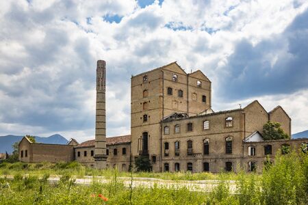 An old disused factory, abandoned and in ruins, with a smashed roof and a chimney. Tall weeds invade...