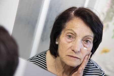 An elderly woman, old lady, looks in the mirror at her aged and wrinkled face, and rubs an anti-wrinkle cream. Dark circles, fatigue, old age, eye bags.の写真素材