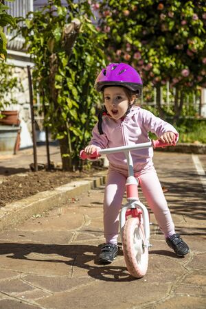 A little girl dressed in pink and wearing a purple helmet plays and enjoys the balance bike. A child runs safely on a bicycle without pedals, in the backyard. Rome Italy.の写真素材