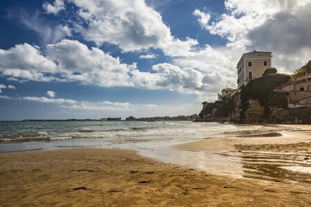 The houses and palaces of the city of Nettuno, on the cliff overlooking the sea. Cloudy blue sky, the wind, the waves of the rough sea. The Roman coast, Lazio, Italy.の写真素材