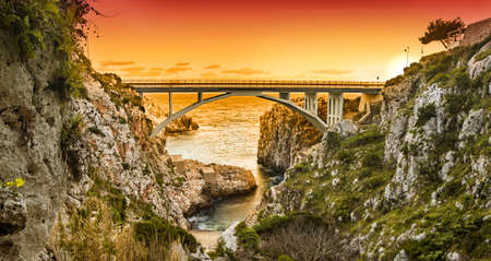 The Ciolo bridge connects two high cliffs. An inlet of the sea, in Gagliano del Capo, near Santa Maria di Leuca. The red and orange sunset. Salento, Puglia, Italy.の写真素材