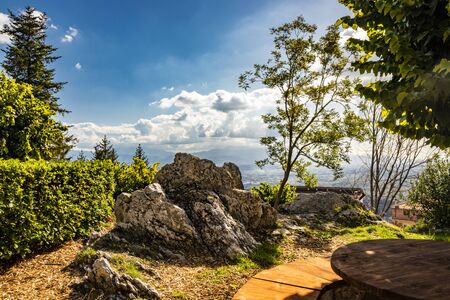 Bellegra, Rome, Lazio, Italy - The panorama seen from Bellegra. The cloudy blue sky. Mountains and sunbeams. Glimpse of a public garden, with trees and spikes of rock. Bench with wooden table.の写真素材