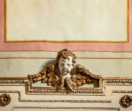 Architectural detail. Bas-relief of a cherub with wings, enriched with stucco friezes, decorations, flowers and frames. Ancient sculpture on a plastered and painted wall.のeditorial素材