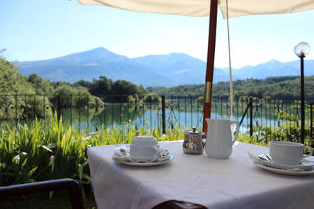 The tables of a hotel for breakfast by the lake. View of the Scandarello lake, in Amatrice, in the province of Rieti, in Lazio, Italy. The blue sky in summer. the green mountains in the background.のeditorial素材