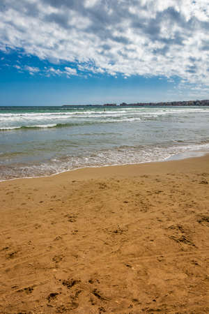 Nettuno, Lazio, Rome, Italy. Some people walk, play and relax on the shore, on the beach of the Roman sea. Backlit. Reflections of light on the water. Cloudy blue sky at sunset. Houses of the city.の写真素材