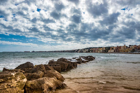 Nettuno, Lazio, Rome, Italy. Some people walk, play and relax on the shore, on the beach of the Roman sea. Backlit. Reflections of light on the water. Cloudy blue sky at sunset. Houses of the city.の写真素材