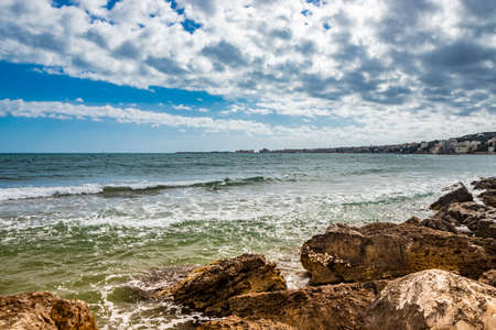 Nettuno, Lazio, Rome, Italy. Some people walk, play and relax on the shore, on the beach of the Roman sea. Backlit. Reflections of light on the water. Cloudy blue sky at sunset. Houses of the city.の写真素材
