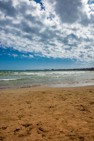 Nettuno, Lazio, Rome, Italy. Some people walk, play and relax on the shore, on the beach of the Roman sea. Backlit. Reflections of light on the water. Cloudy blue sky at sunset. Houses of the city.の写真素材