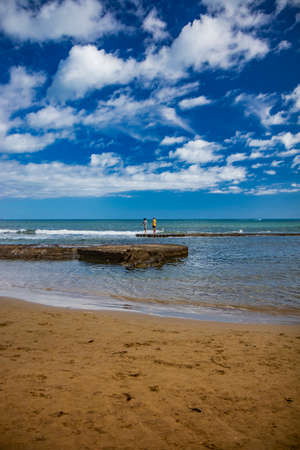 Nettuno, Lazio, Rome, Italy. Some people walk, play and relax on the shore, on the beach of the Roman sea. Backlit. Reflections of light on the water. Cloudy blue sky at sunset. Houses of the city.のeditorial素材