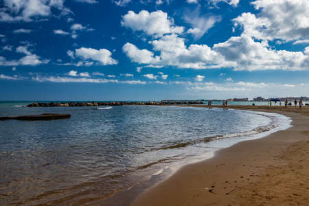 Nettuno, Lazio, Rome, Italy. Some people walk, play and relax on the shore, on the beach of the Roman sea. Backlit. Reflections of light on the water. Cloudy blue sky at sunset. Houses of the city.のeditorial素材
