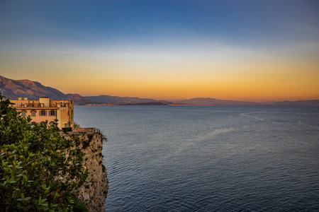 Gaeta, Latina, Lazio, Italy. A beautiful sunset over the sea, seen from a panoramic viewpoint in the village. A building overlooking the sea, perched on a sheer cliff. Mountains in the background.の写真素材