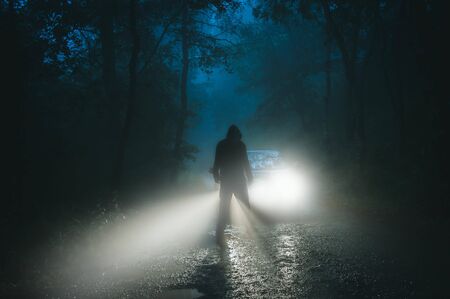 A sinister hooded figure standing in front of a car. On a spooky forest road on a foggy evening.の写真素材