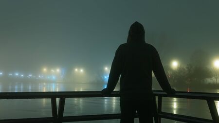 A hooded figure silhouetted against street lights next to a river. On a mysterious misty, winters nightの写真素材